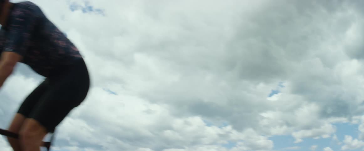 A man wearing a helmet is riding a bicycle on a road overlooking mountains under a cloudy sky