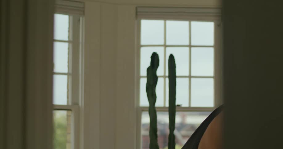 A man is standing in a room getting dressed as he pulls an undergarment over his shoulders and puts on a black shirt