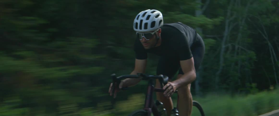 A cyclist is leaning over his handlebars as he coasts down a hill on a road through the woods