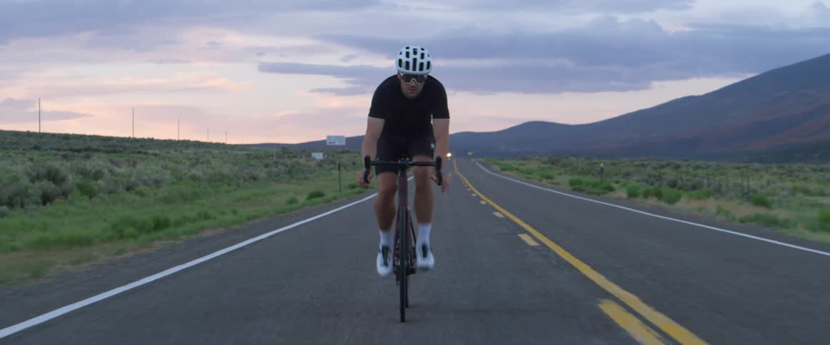 Man wearing shorts and sunglasses rides a bicycle on a highway next to the field with mountains in the distance