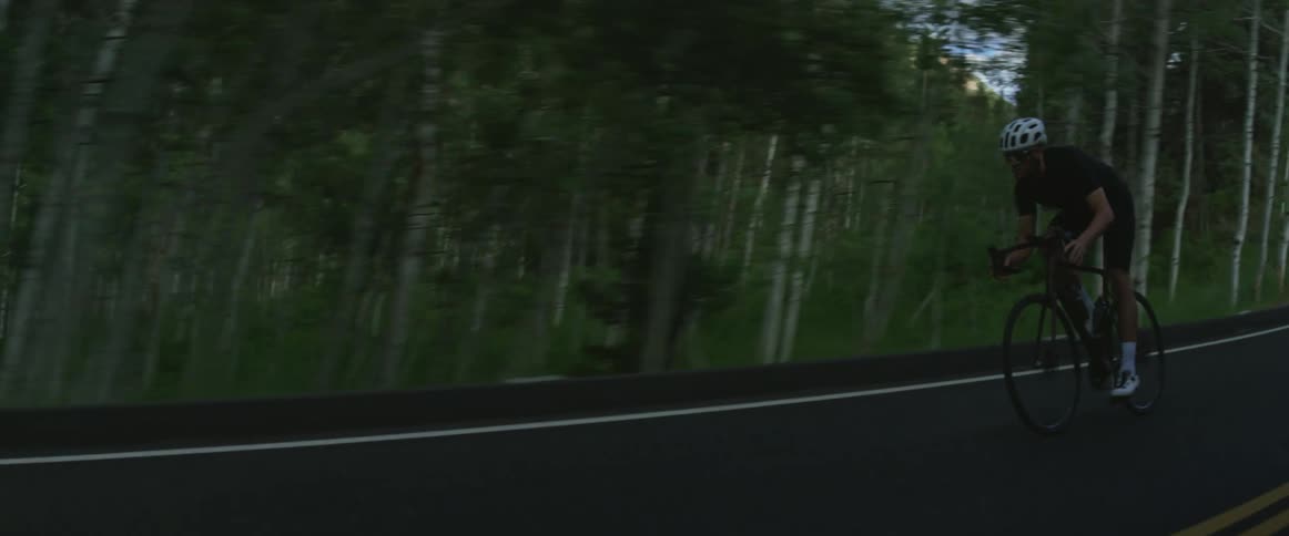 Man wearing sunglasses and a helmet rides a bike on a highway next to a forest