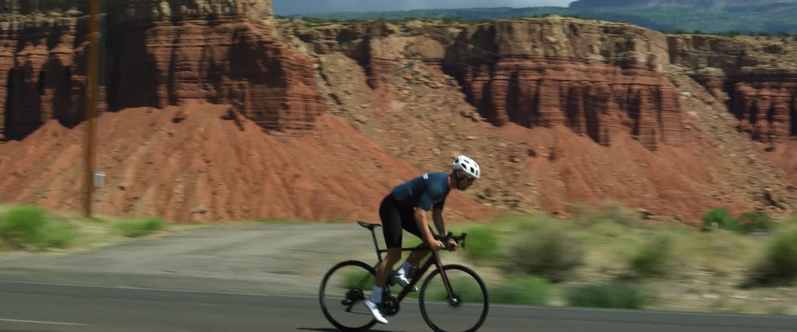 Man wearing a helmet and sunglasses and shorts rides a bike on a rural highway with a mesa nearby