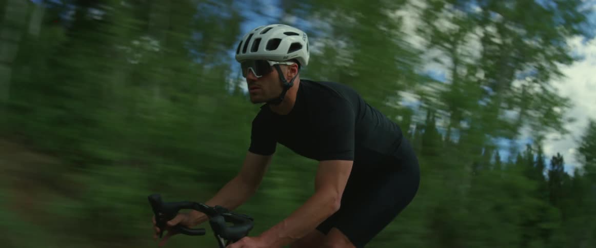 Man wearing a helmet and sunglasses rides a bicycle next to a forest under a sunny blue sky
