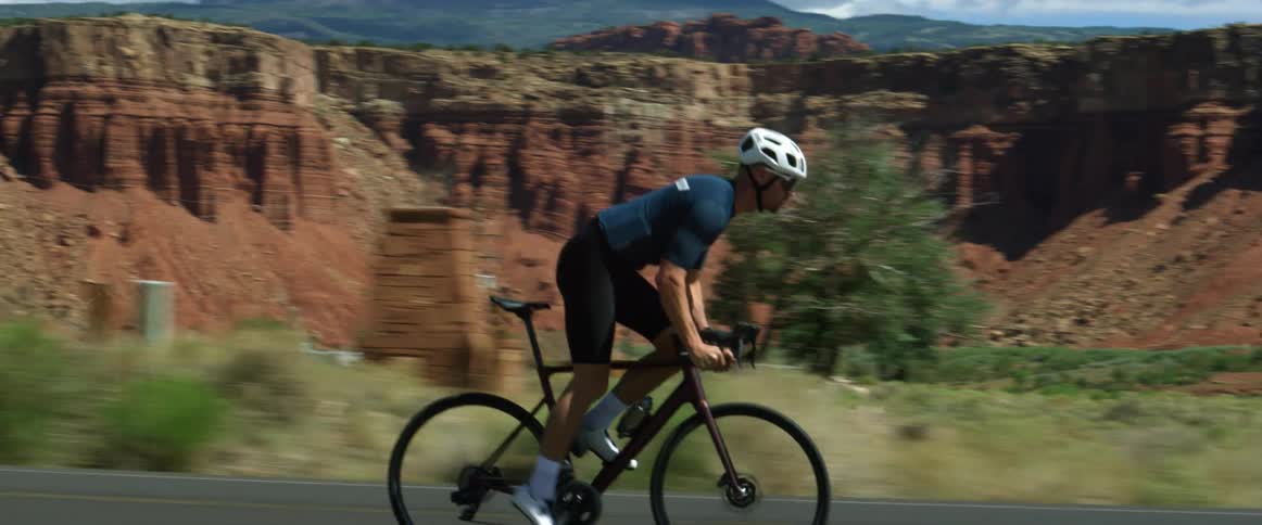 A man wearing a helmet is pedaling hard as he rides a bicycle on a road by rock formations and badlands