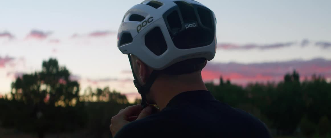A man places a bicycle helmet on his head and fastens the strap while outdoors at sunset