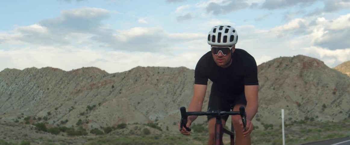 Man wearing sunglasses and a helmet rides a bicycle on a highway near the mountains on a sunny day