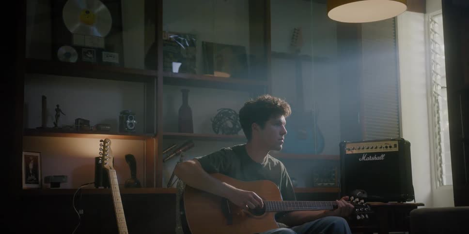 Teenaged boy sits and plays the guitar in a room with shelves on a wall and blinds on the window