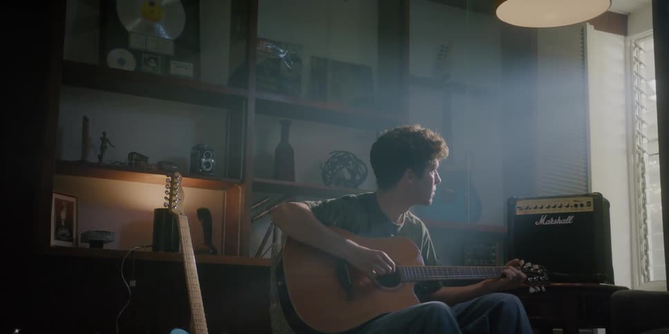 A young man plays a guitar while sitting indoors and gazing out the window during the day