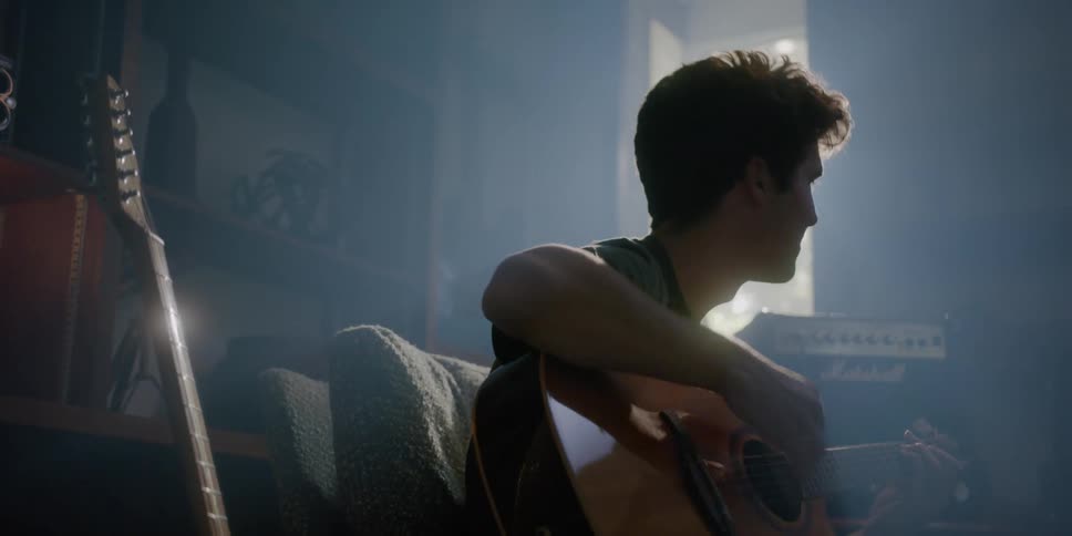 Teenaged boy plays the guitar while sitting on a chair in front of some shelves