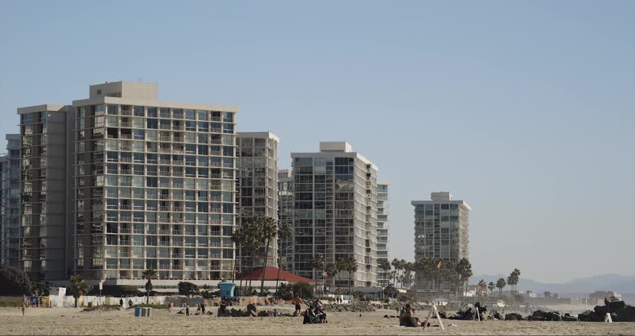 Birds fly through air as people move around beach near apartment buildings with mountains on the horizon