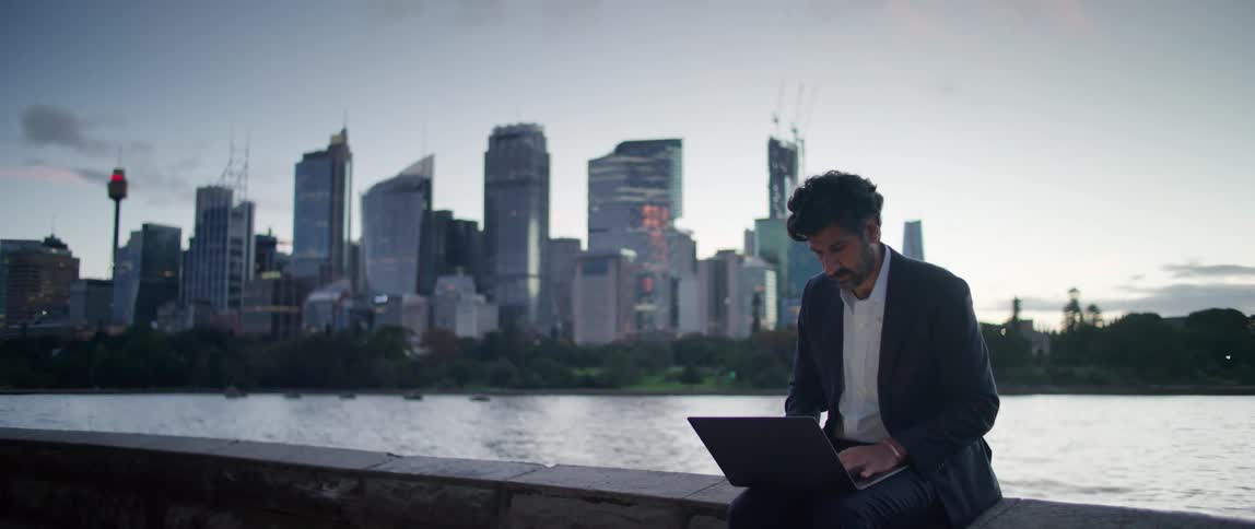 A man is sitting by the lakeside working on a laptop computer and there is a big city with towers and skyscrapers in the distance