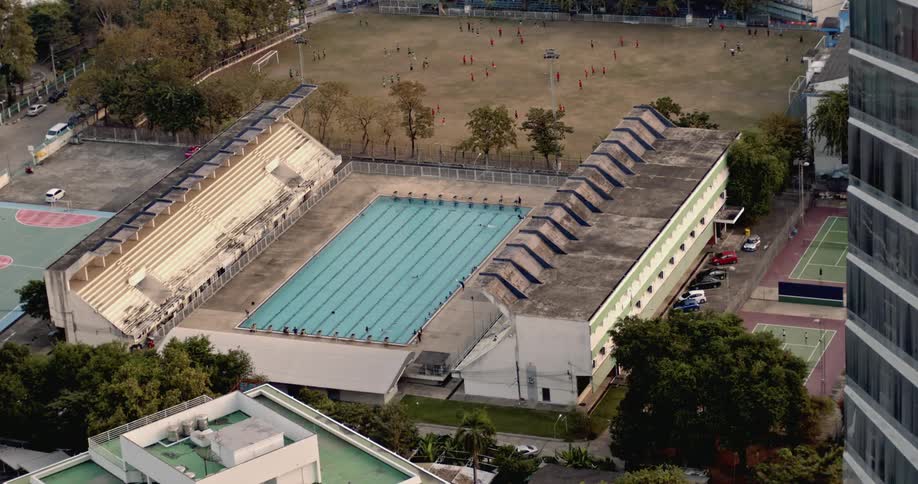 A long swimming pool can be seen from high above next to some buildings and a soccer field
