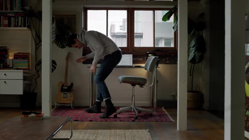 A man puts a guitar down then sits at a desk and uses a laptop