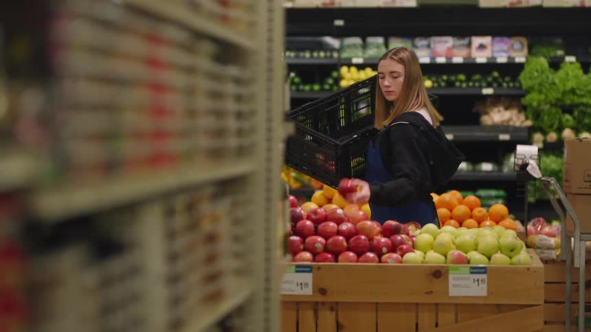 Young girl working at a small supermarket picking up apples from a stand