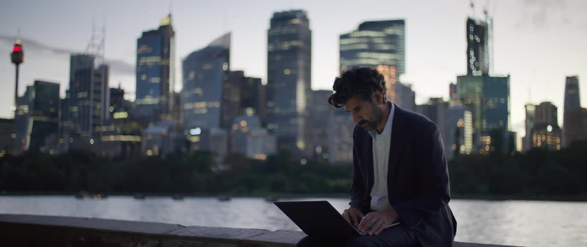 A man is sitting near a river that flows through a city as he types on his laptop