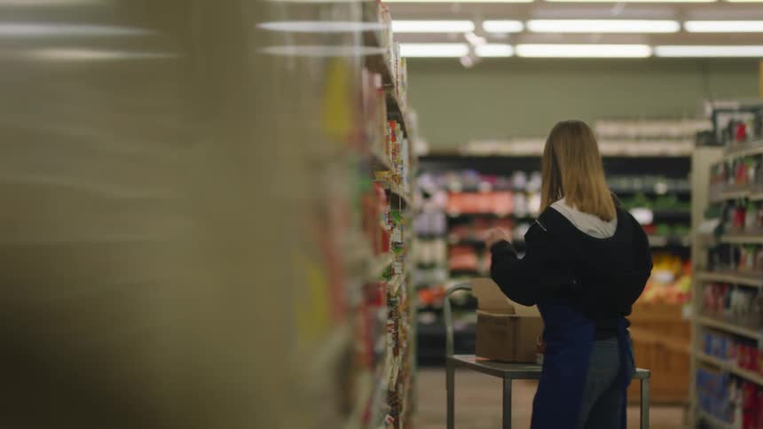 A woman is stocking cans on a shelf at a store as another worker walks in another aisle