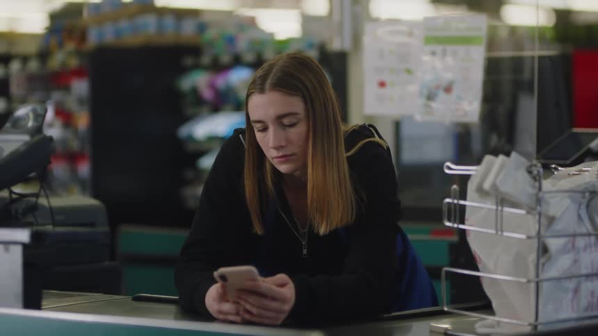 Young bored woman looks at her phone while working at a small supermarket
