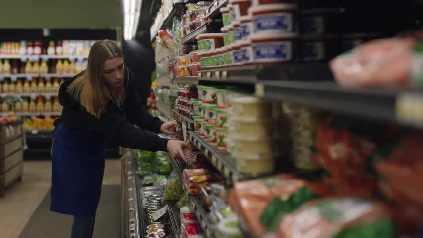A woman is fixing signs and straightening up the produce on a shelf in a store