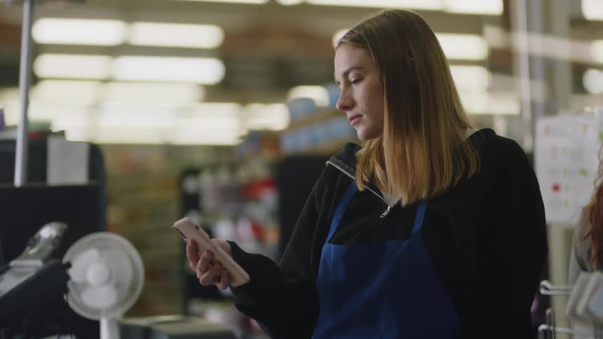 A cashier looks bored as she stands in a store and looks at her cell phone