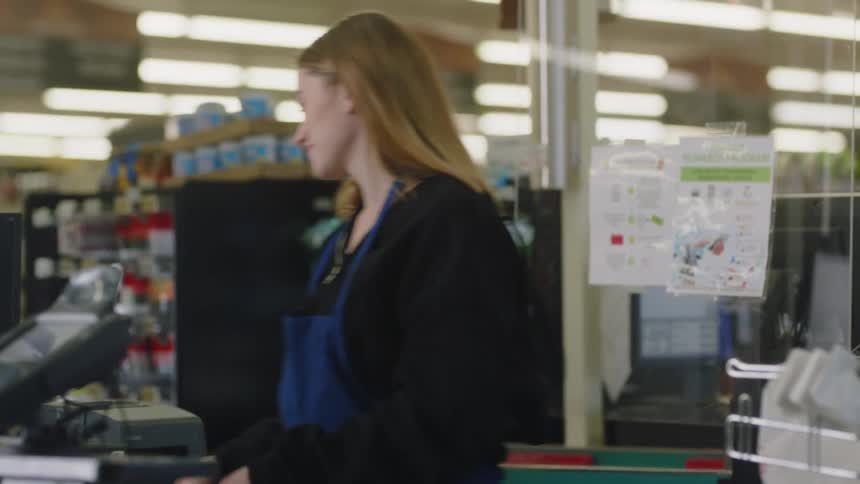 A teenage girl working as a cashier takes her headphones out and smiles