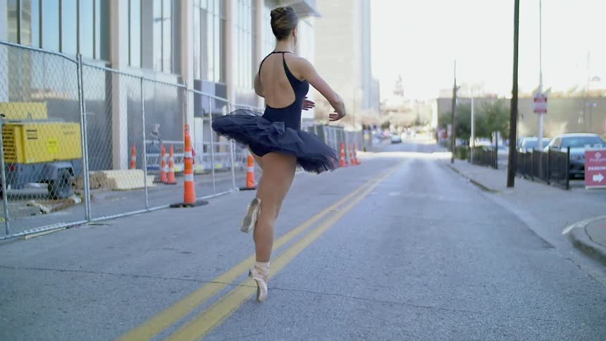 A ballerina dances on a city street near a chain link fence