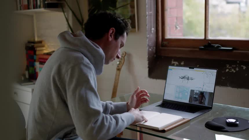 A man sits at a desk in front of his laptop while having a discussion via video meeting