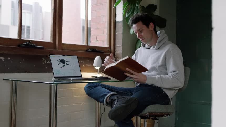 A man is reading a book and looking on a computer as he sits at a desk