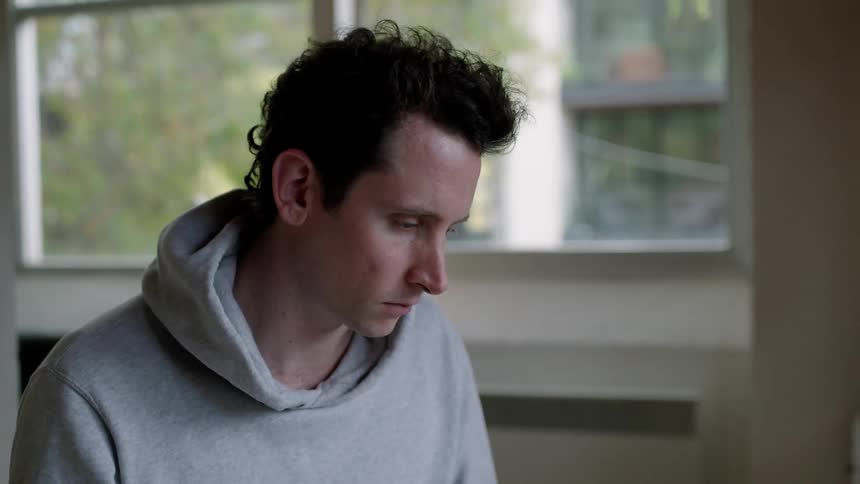A man is sitting at a desk talking to someone on a computer in front of a window