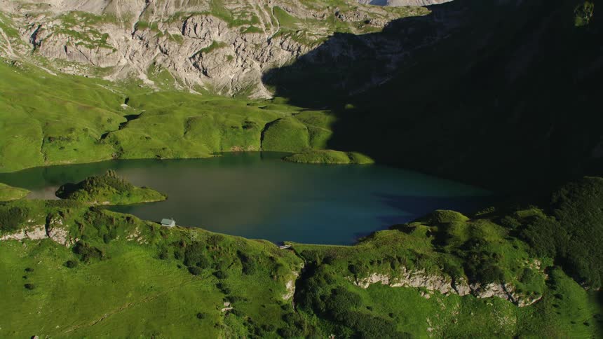 A small lake is nestled high up within a mountain range that is covered with greenery
