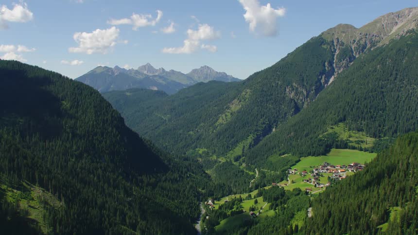 Forested mountains with a village on a mountainside plateau and mountains in the distance under a cloudy sky