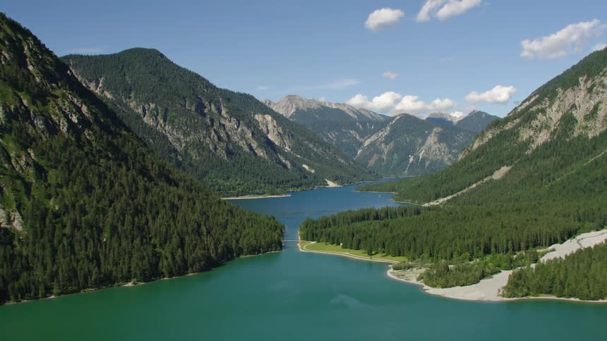 A bridge crosses a blue lake with forests along the shore and surrounded by mountains with cumulus clouds in the sky
