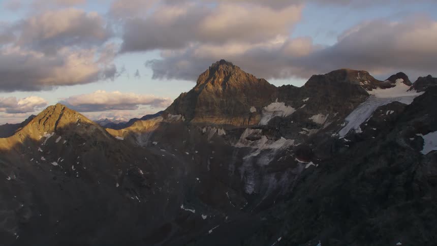 The sun shines on mountains as clouds hoover over the peaks and ridges