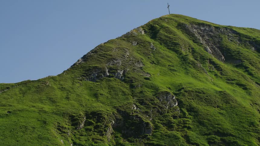 A grass covered mountain is high in a cloudy sky with a cross on the peak