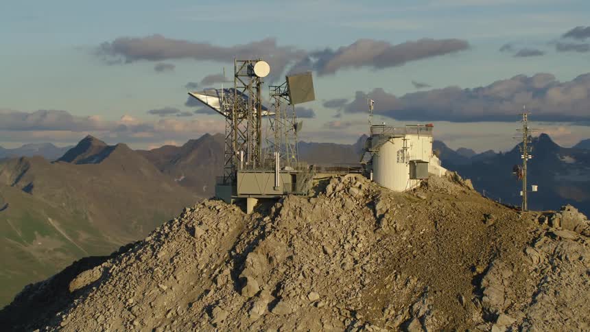 A radar tower is high on the Valluga mountaintop and there are mountains in the distance under a cloudy sky