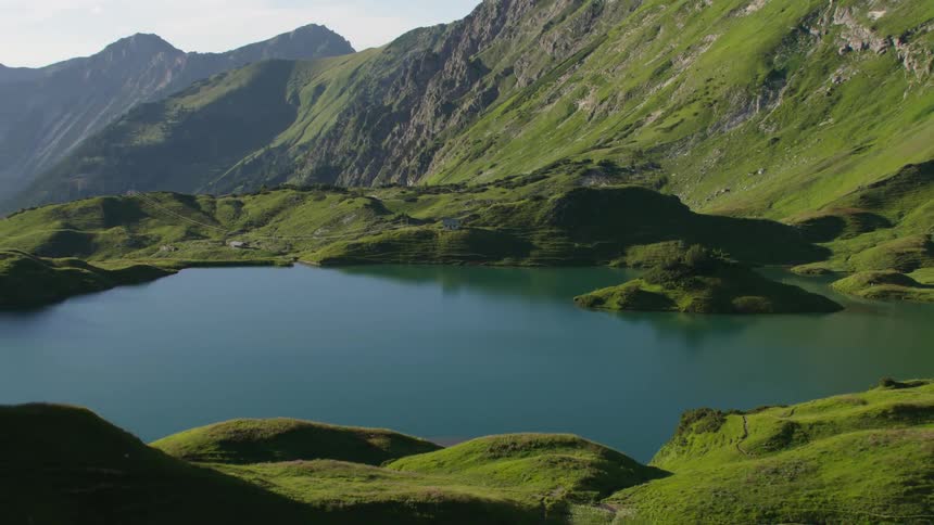 Grass covered land surrounds a lake at the foot of a mountain and there are mountains in the distance under a cloudy sky