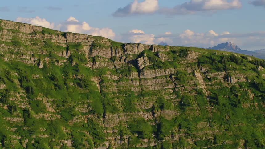 Densely vegetation covered cliff side stands under a cloudy sky with other mountains in the background