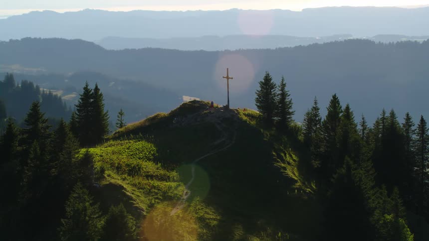 A person is standing on a forested mountaintop near a cross and there are mountains in the distance