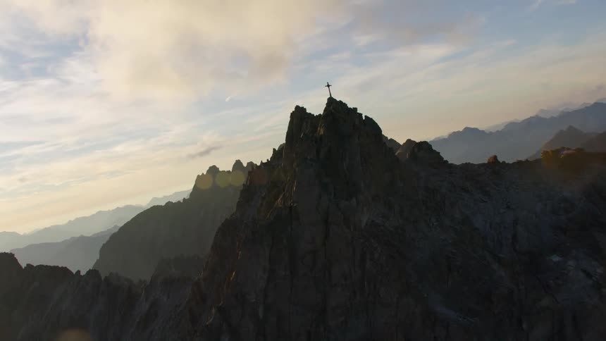 The camera twists as it flies over a cross thats placed atop a mountain summit