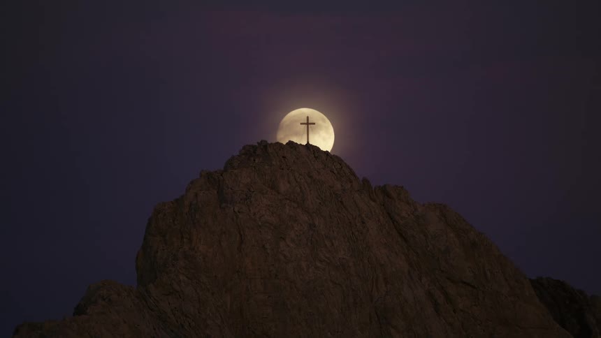 A cross atop a mountain summit is back lit by the moon