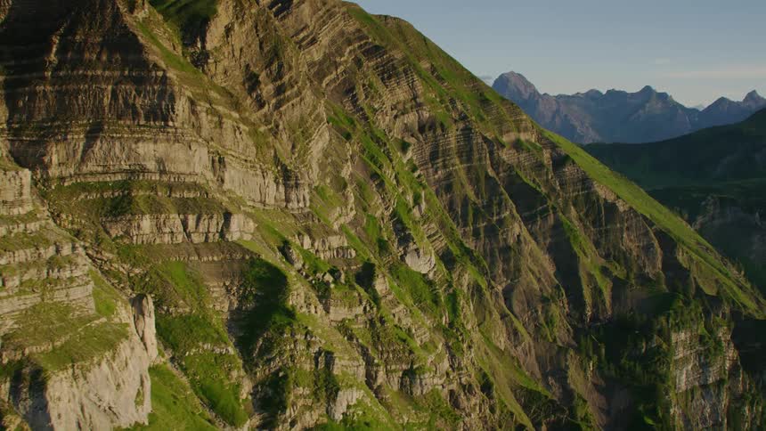 A towering mountain range has an impressive sloping cliff side that is covered in vegetation