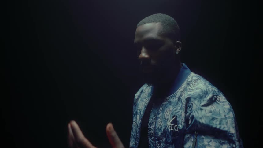 A black man is standing in the darkness and he looks at his left hand and raises it in the air