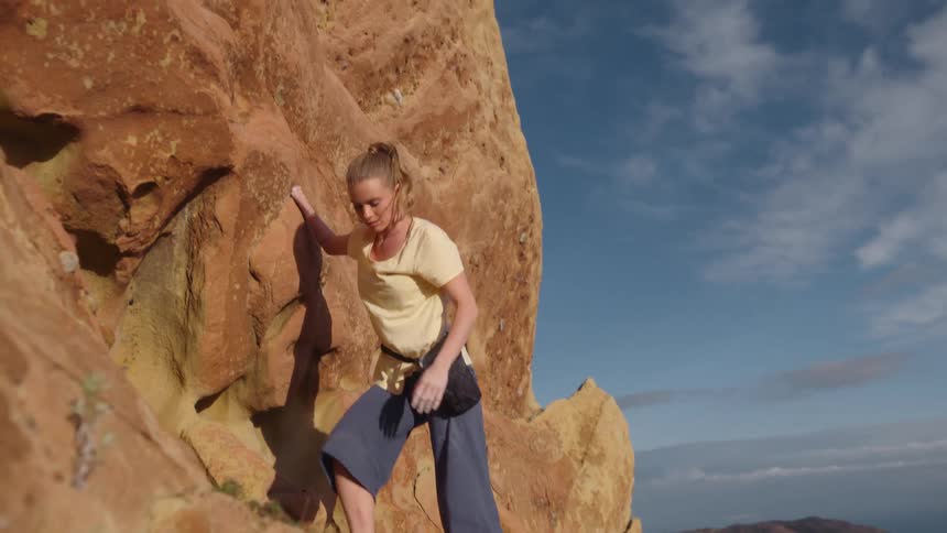 A woman is climbing up the side of a mountain and standing on a ledge as she reaches above her head