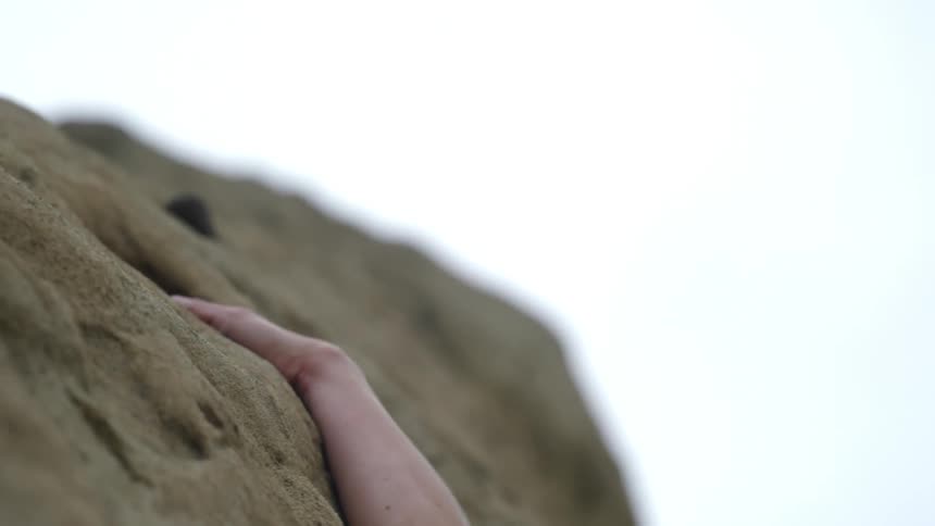 Blonde woman with a pony tail reaches with her arm to pull herself up the rocks on the side of the mountain