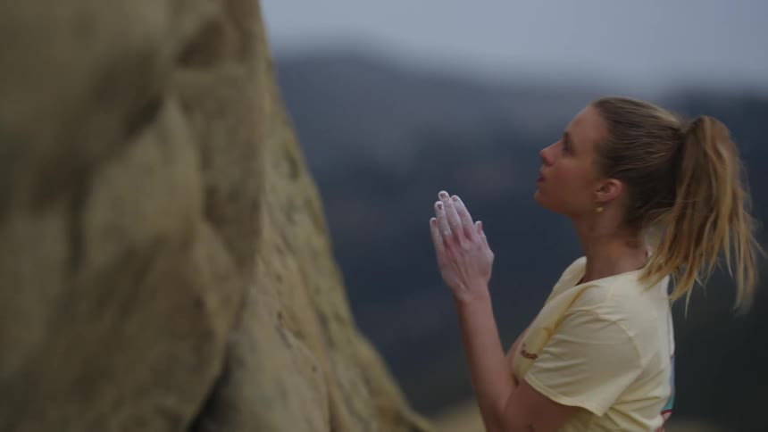 A woman mountain climbing is standing on a ledge reaches into her pouch and rubs powder on her hands