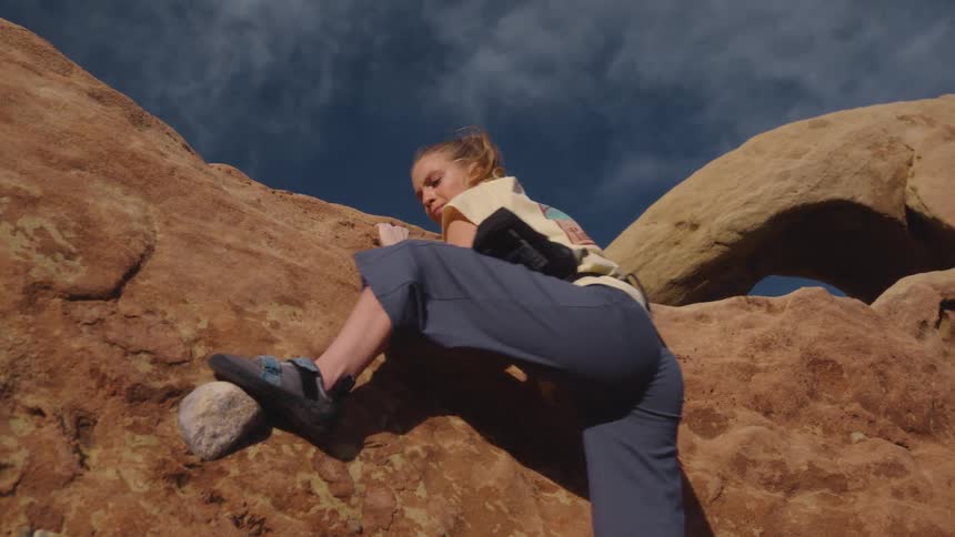 A woman climbing up the side of a mountain lifts her leg on to a rock