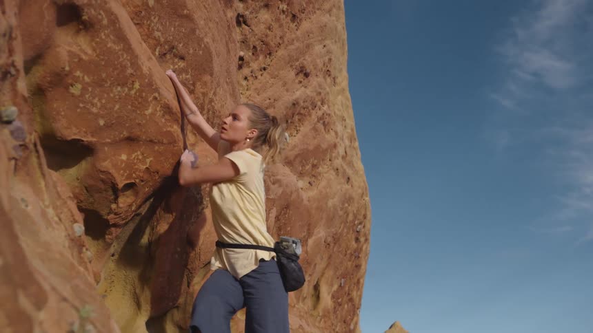 A woman is climbing up the side of a mountain and holding on to rocks as she secures her footing