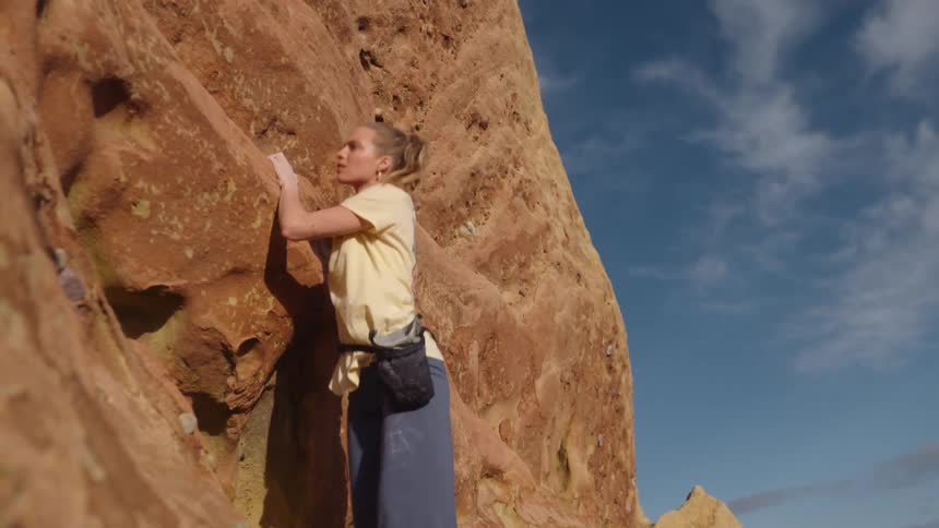 A woman is climbing up the side of a mountain and holding on to rocks as she moves across a ledge