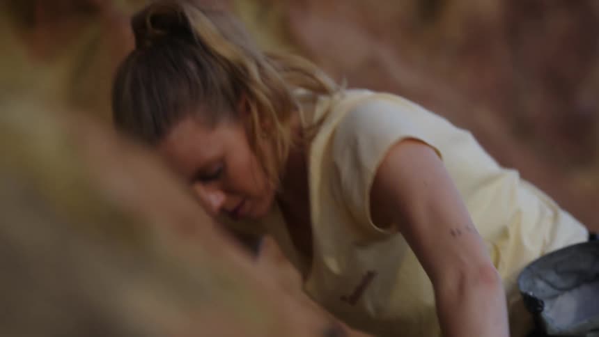 A woman is climbing the side of a mountain and holding on to rocks as she pulls herself up to a ledge