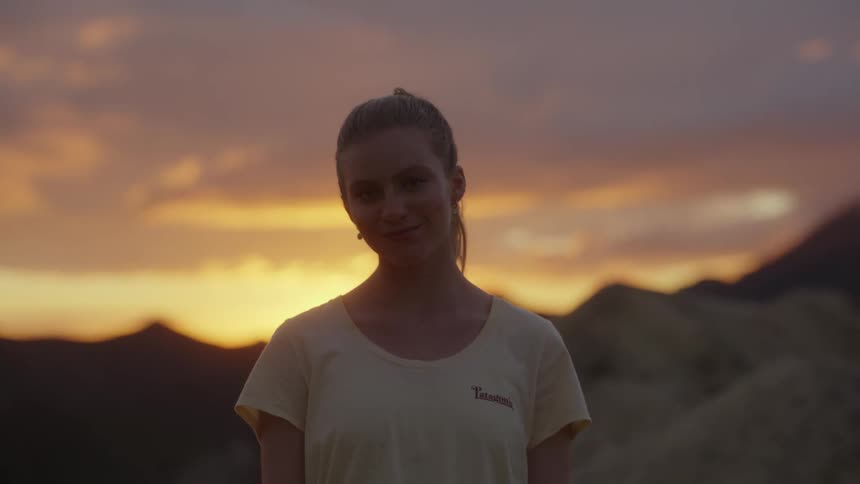 A woman stands outdoors as the sun is setting and clouds drift in the sky behind her
