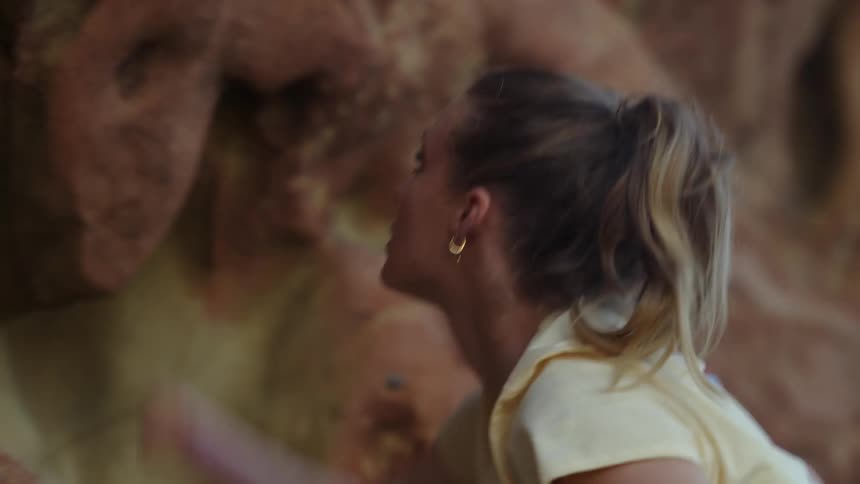 A woman is climbing the side of a mountain and looks up as she grabs ahold of rocks above her head